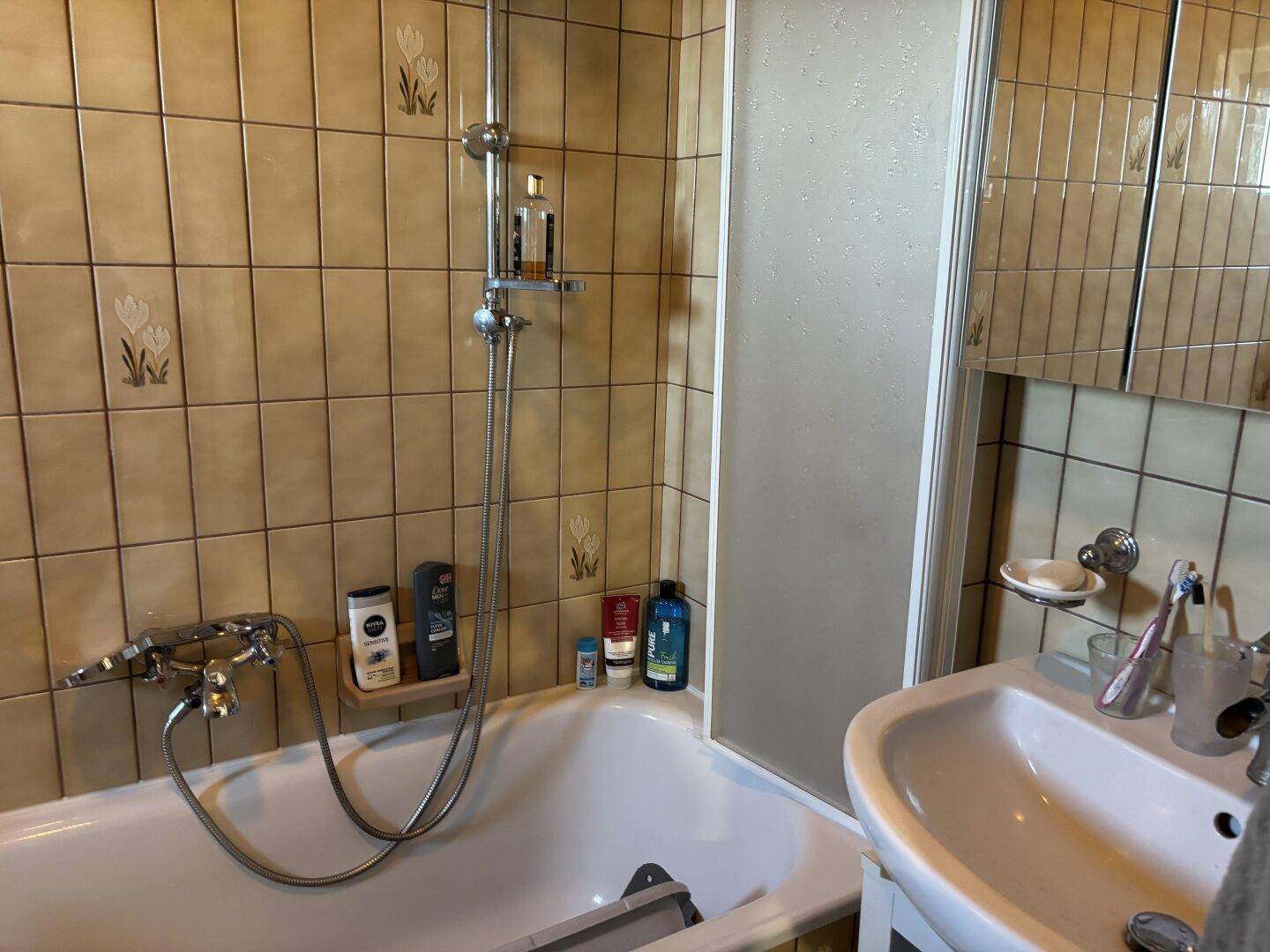Badezimmer mit Badewanne, Duschabtrennung und beigen Fliesen im Stil der 80er Jahre.