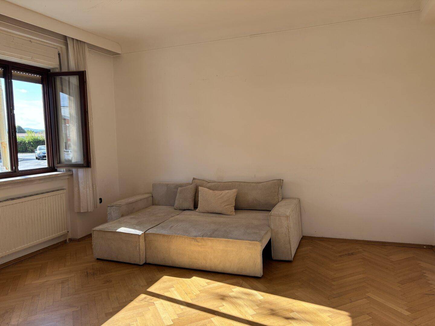 Helles Wohnzimmer mit einem beigen Sofa, Fischgrätparkett und einem geöffneten Fenster.