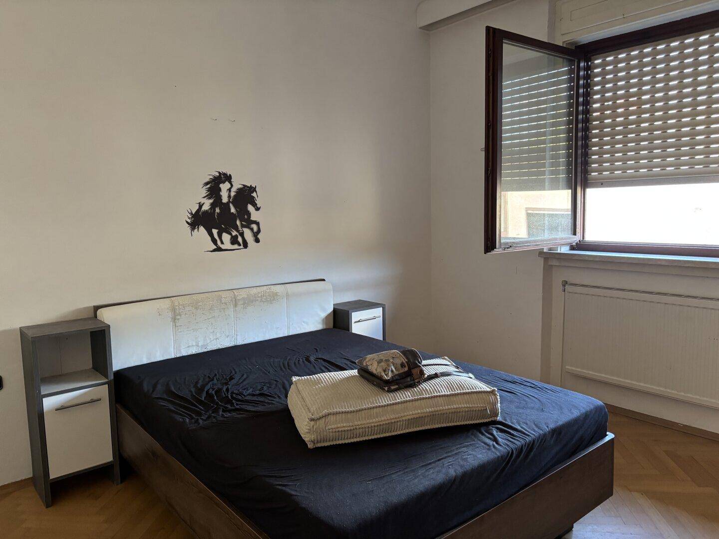 Schlafzimmer mit Doppelbett, Nachttischen und einem geöffneten Fenster mit Rollladen.