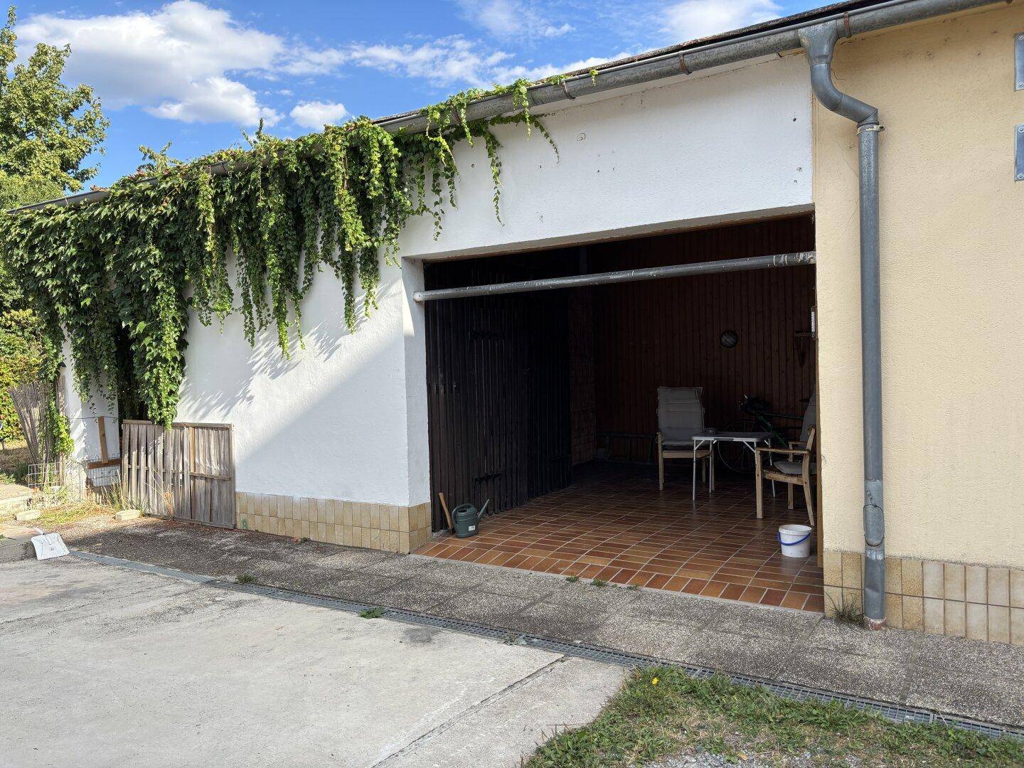Geräumige Garage mit Fliesenboden und Efeu bewachsener Fassade, die auch als überdachter Sitzbereich genutzt werden kann.