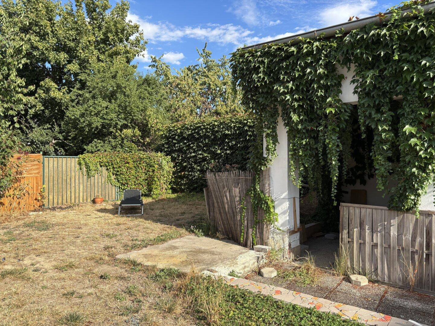 Trockener Gartenbereich mit Hecke, Bäumen und einem Liegestuhl, umgeben von einem Holzzaun.