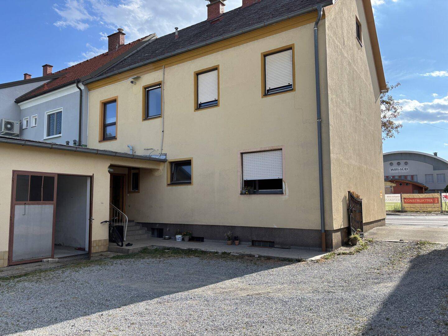 Seitenansicht des gelben Wohngebäudes mit Garage und Schotterauffahrt, umgeben von weiteren Gebäuden.