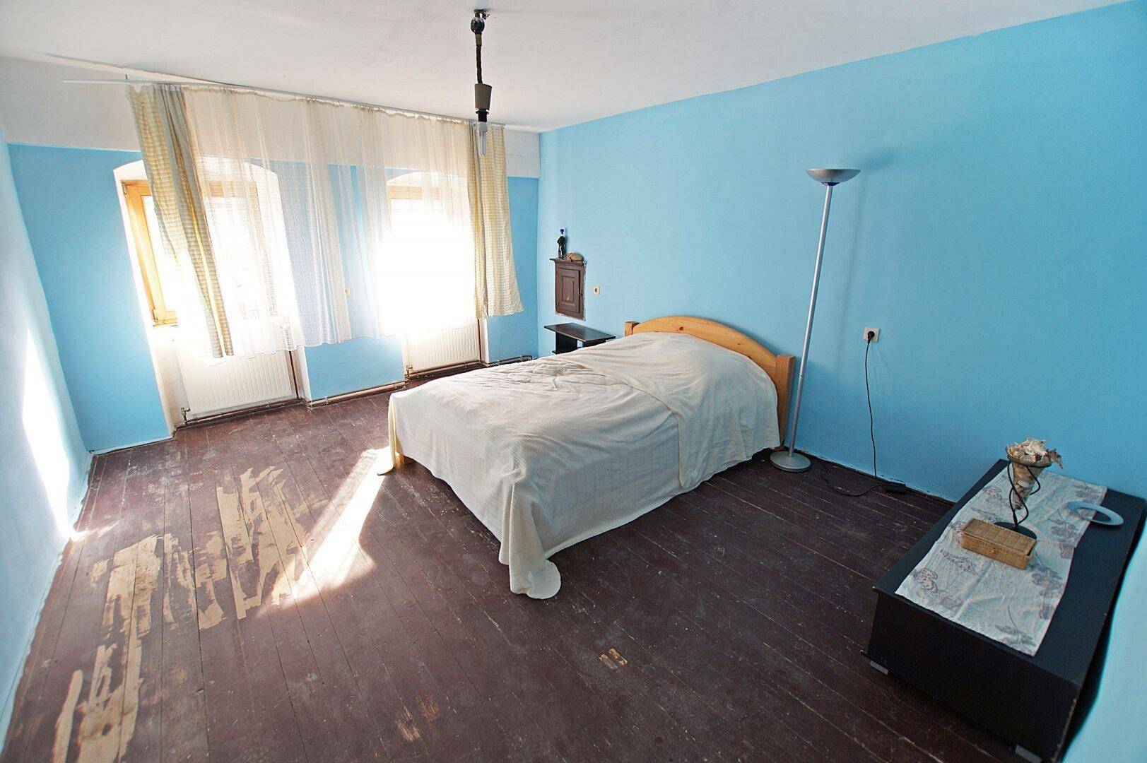 Schlafzimmer mit blauen Wänden, Holzdielenboden und viel Tageslicht durch die Fenster.