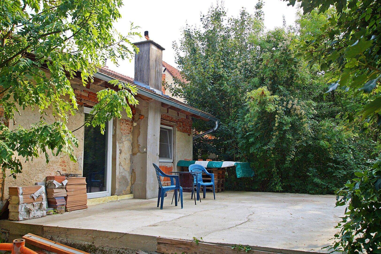 Betonierte Terrasse am Haus mit Gartenmöbeln und umgeben von Bäumen und viel Grün.