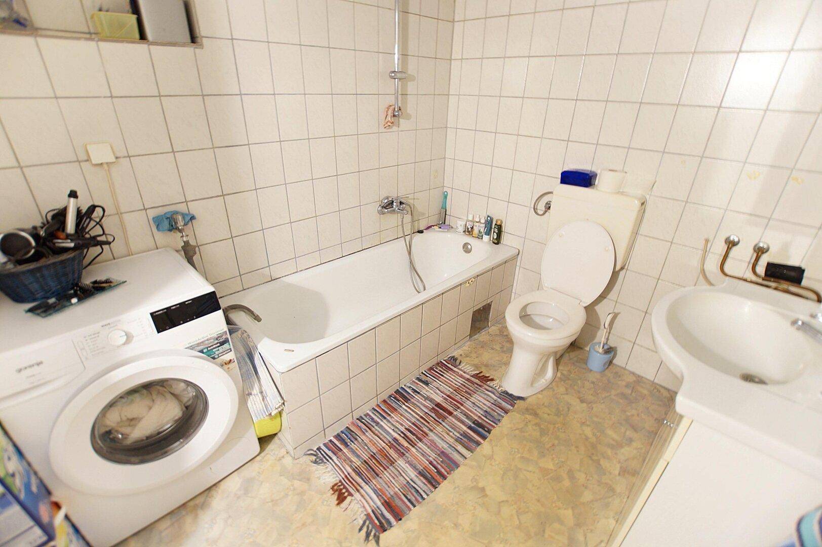 Badezimmer mit Badewanne, Waschmaschine und weißen Fliesen an den Wänden und auf dem Boden.