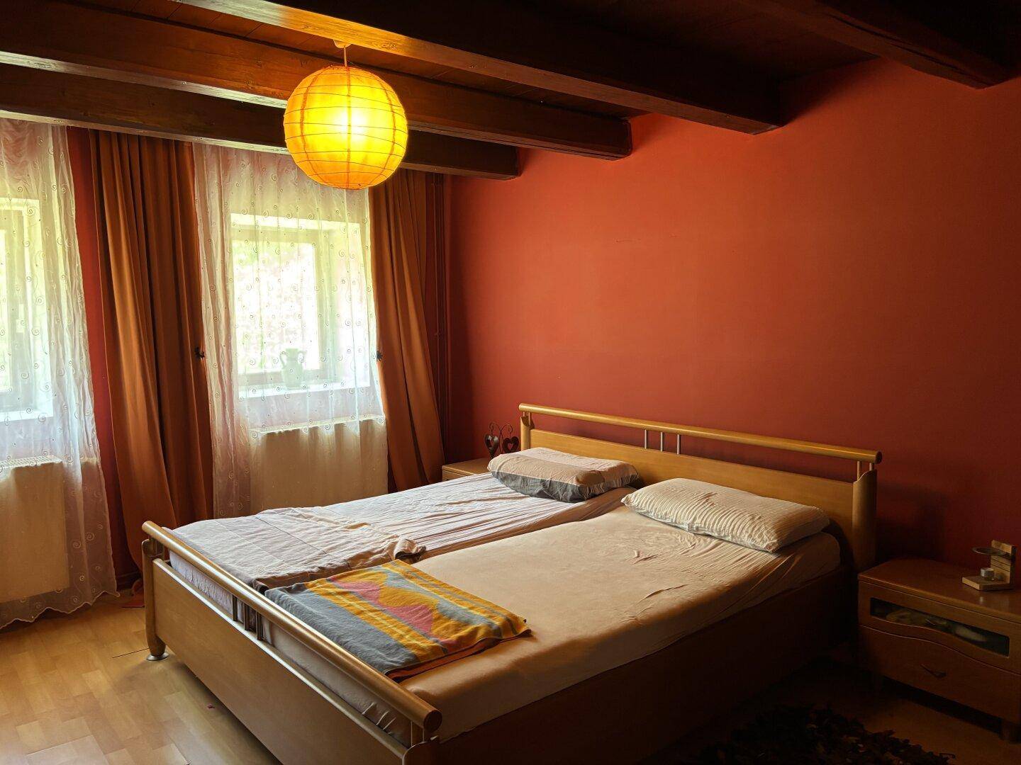 Schlafzimmer mit Holzbalken an der Decke, roter Wand und zwei Fenstern für natürliches Licht.