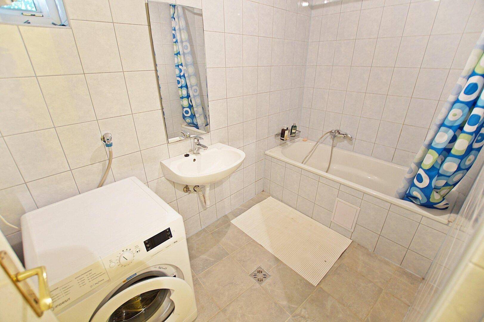 Badezimmer mit Badewanne, Waschmaschine und weißen Fliesen an den Wänden und auf dem Boden.