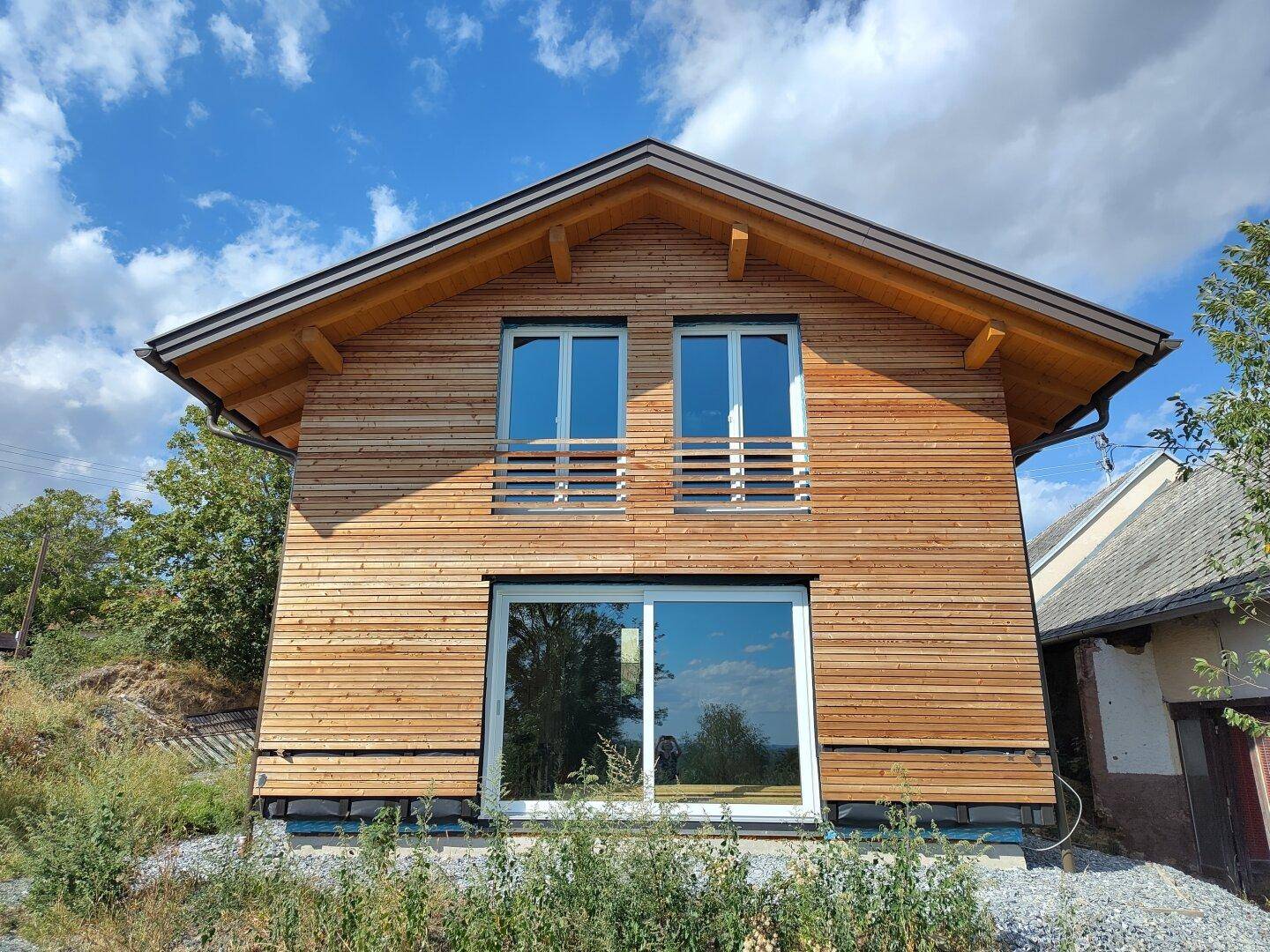 Frontansicht des modernen Holzhauses mit großen Fenstern und Holzbalkon-Geländern unter blauem Himmel.