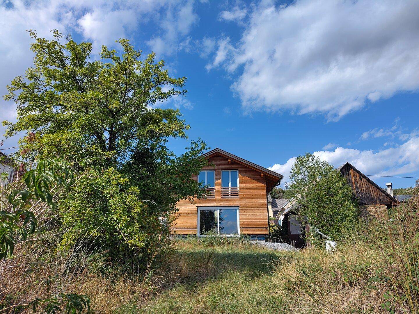 Holzhaus eingebettet in grüne Natur mit Bäumen und Sträuchern unter blauem Himmel.