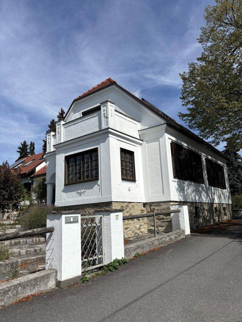 Weißes Gebäude mit rotem Ziegeldach und dekorativen Fassadenelementen an einer leicht ansteigenden Straße.