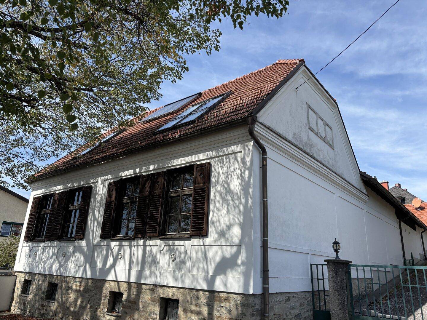 Traditionelles Haus mit weißer Fassade, braunen Fensterläden und Ziegeldach mit Dachfenstern.