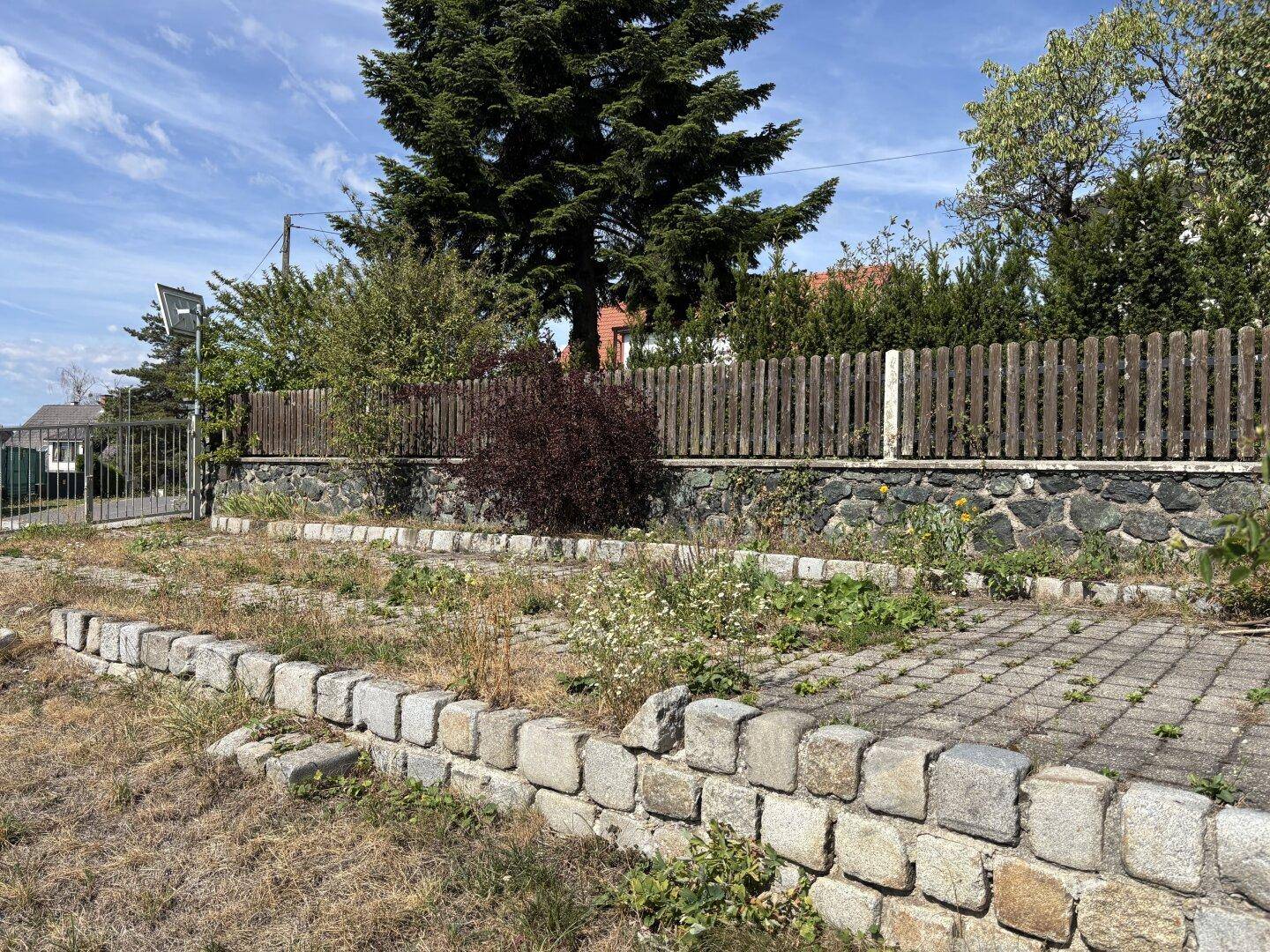 Vernachlässigter Gartenbereich mit Steinmauern und Holzzaun, der auf Pflege wartet.