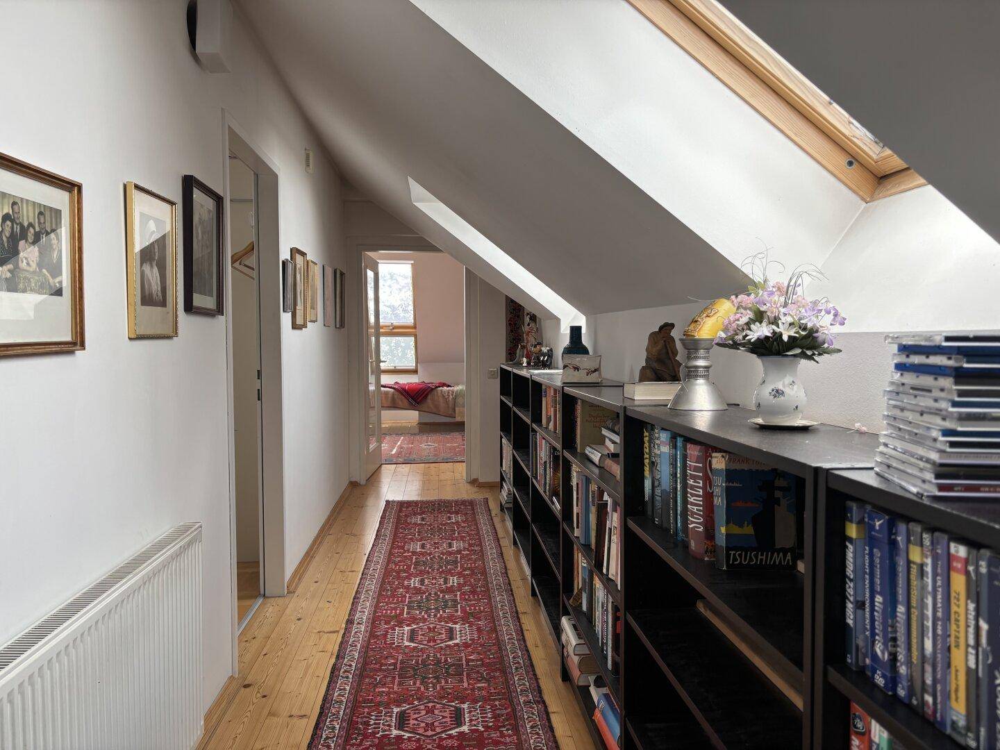 Heller Dachgeschossflur mit Holzboden, Bücherregalen und Dachfenstern, der zu weiteren Räumen führt.
