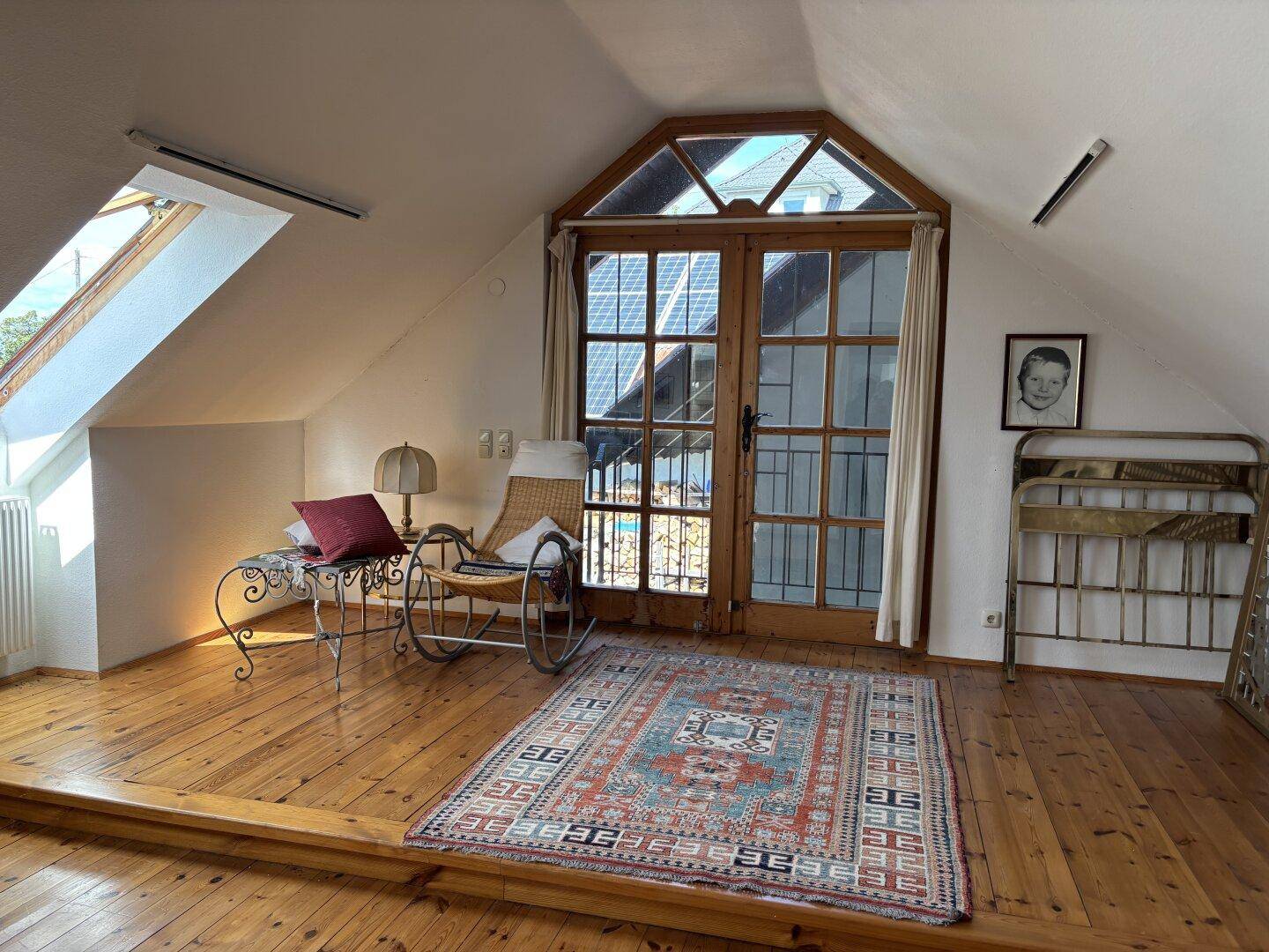 Helles Dachzimmer mit Holzboden, Schaukelstuhl und Zugang zu einem französischen Balkon mit Blick ins Grüne.