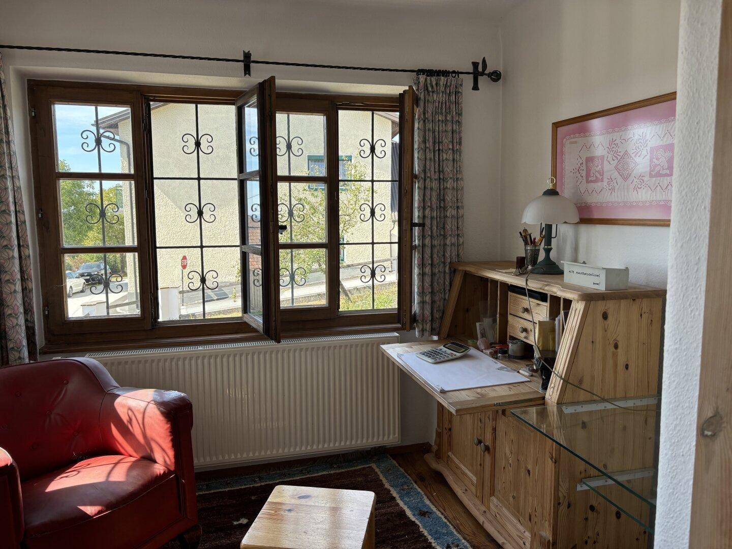 Helles Arbeitszimmer mit Holzmöbeln, rotem Sessel und Blick ins Grüne durch Sprossenfenster.