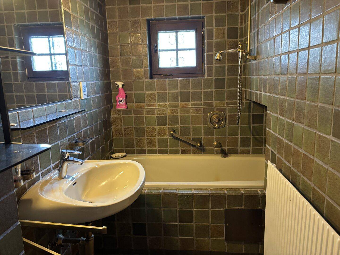 Badezimmer mit grünen Fliesen, Waschbecken, Badewanne mit Duschfunktion und kleinem Fenster.