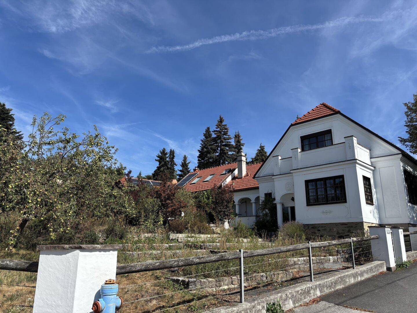 Historisches weißes Gebäude mit rotem Ziegeldach und großem, naturbelassenem Gartenbereich unter blauem Himmel.