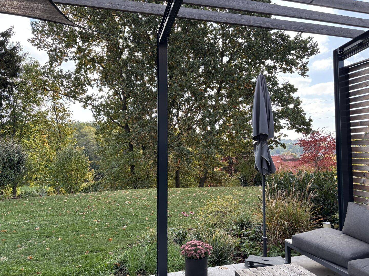 Gemütlicher Außenbereich unter einer Pergola mit Blick auf den weitläufigen Garten und die umliegende Natur.