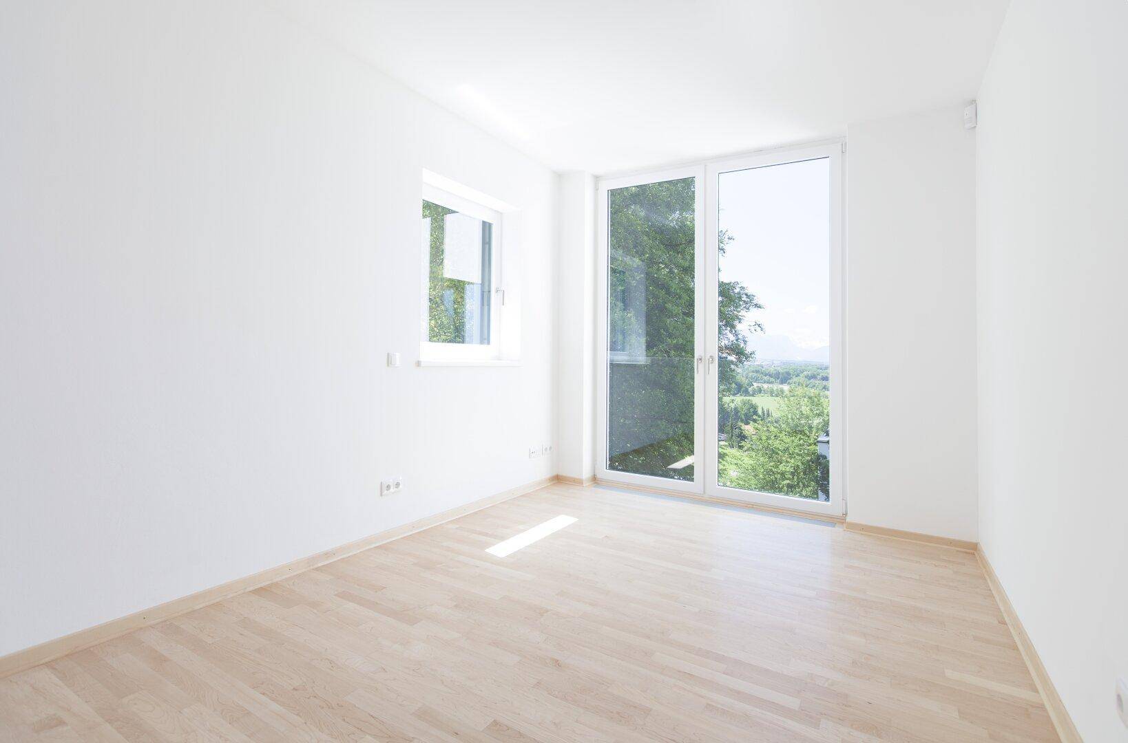 Lichtdurchflutetes Zimmer mit Holzboden und Blick ins Grüne durch große Fenster und Balkonzugang.