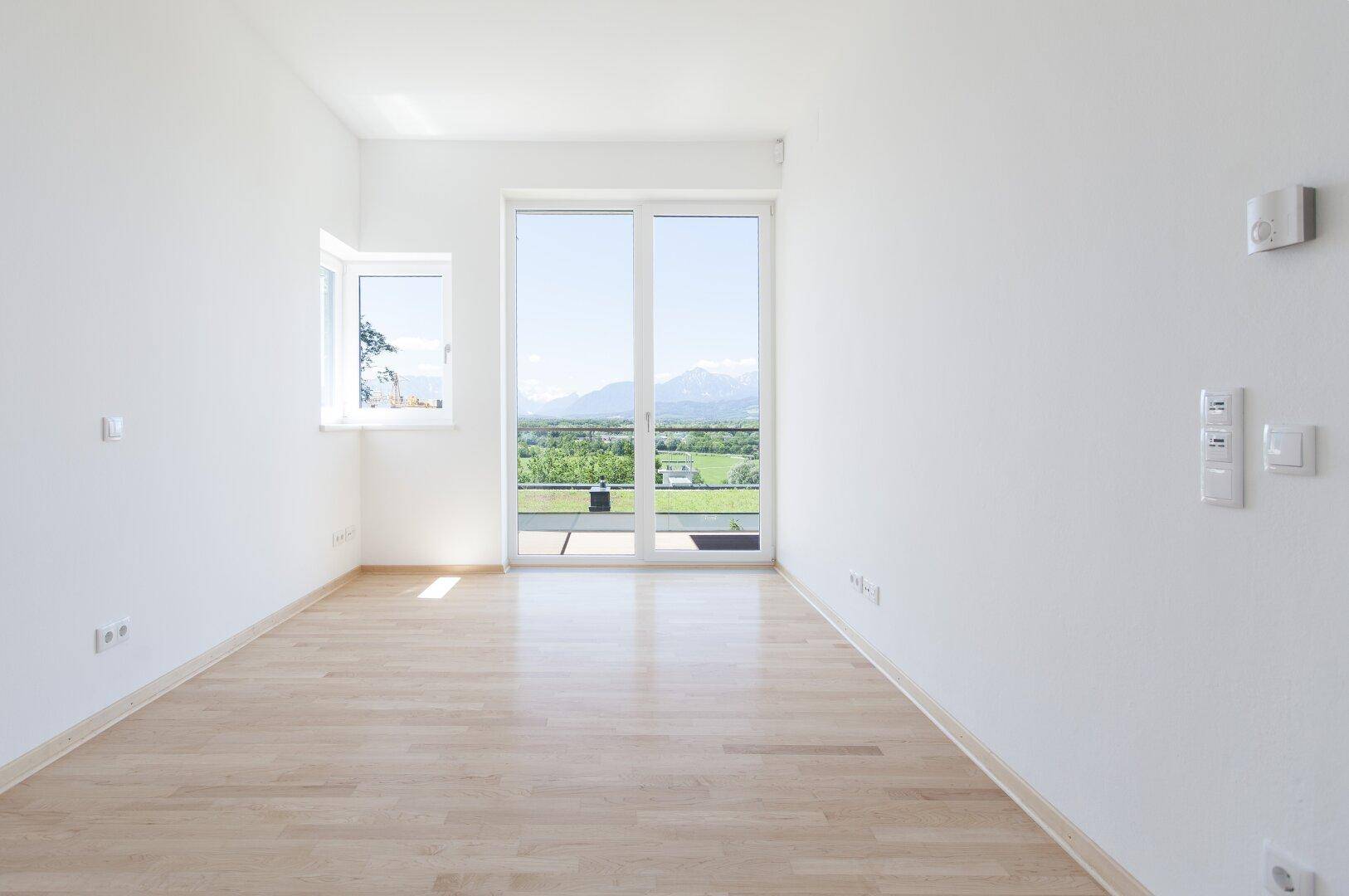 Helles Zimmer mit Holzboden, großen Fenstern und weitem Blick auf die umliegende Landschaft und Berge.