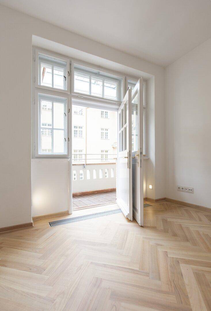 Heller Raum mit Fischgrätparkett und geöffneter Balkontür, die einen Blick auf die Umgebung freigibt.
