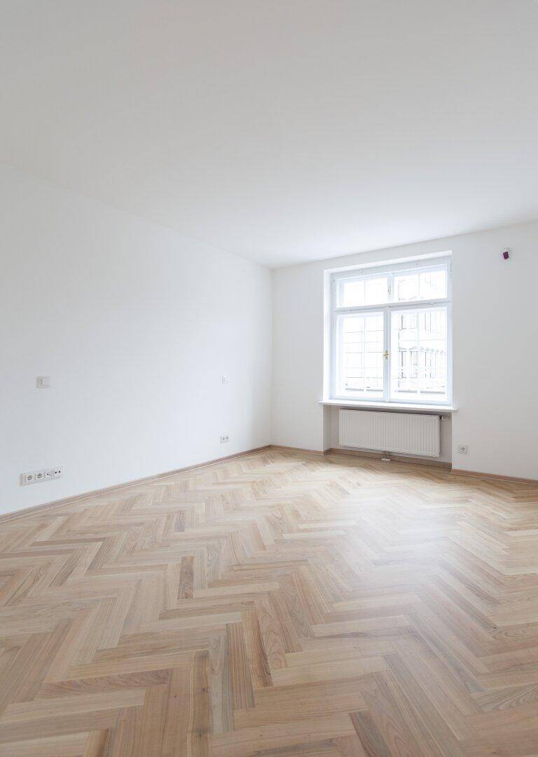 Leerer Raum mit Fischgrätparkett und einem großen Fenster, das viel Tageslicht hereinlässt.