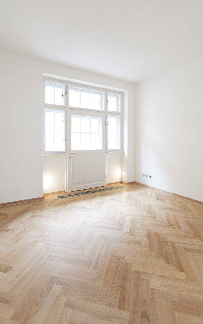 Heller Raum mit Fischgrätparkett und einer Balkontür, die viel Tageslicht hereinlässt.