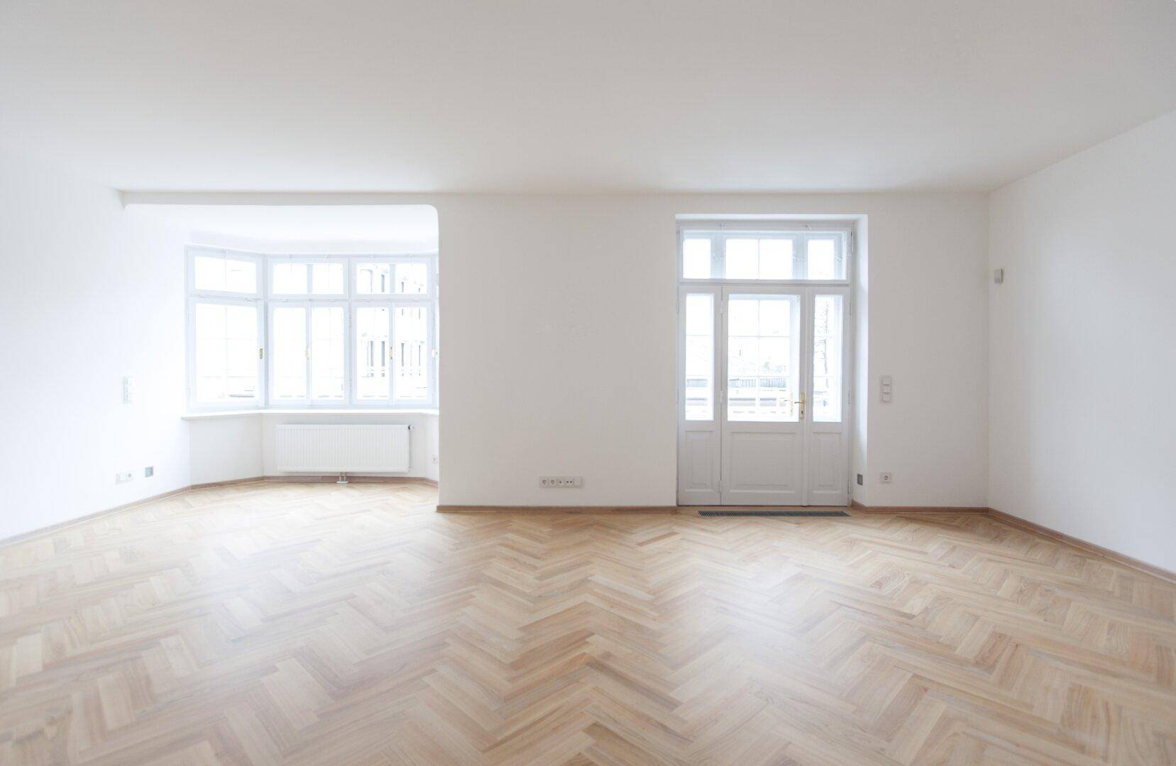 Großes, helles Wohnzimmer mit Fischgrätparkett, Erkerfenstern und Zugang zu einem Balkon.