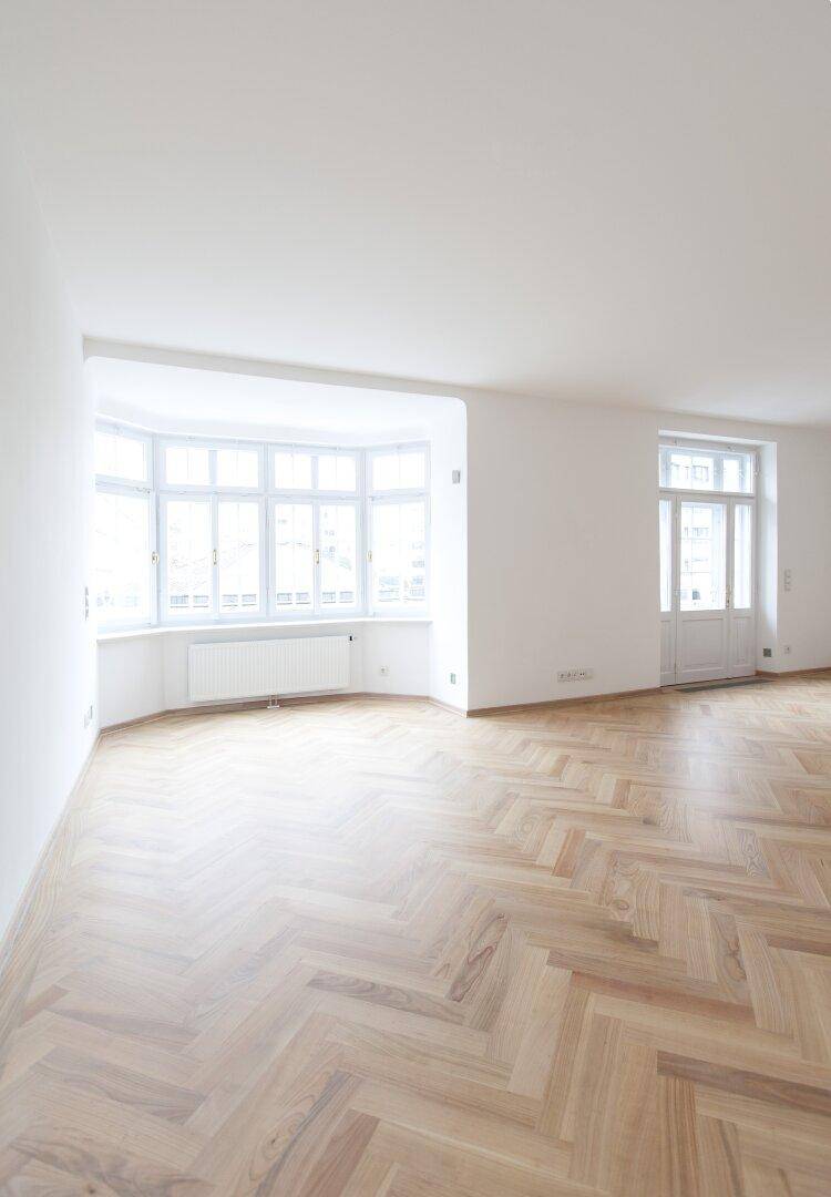 Geräumiges Wohnzimmer mit hellem Fischgrätparkett und einem großen Erkerfenster, das viel Licht hereinlässt.