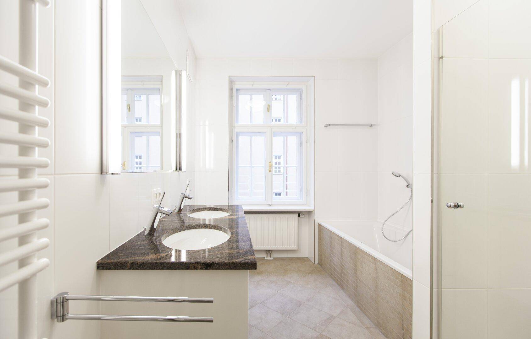 Helles Badezimmer mit Doppelwaschbecken, Badewanne, Dusche und Fenster.