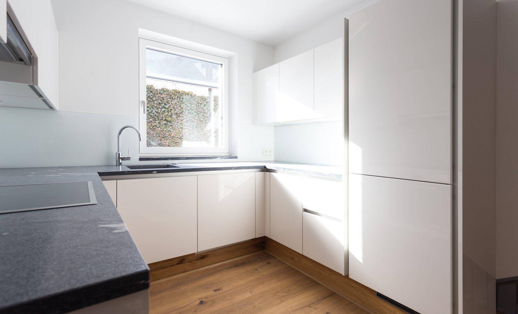Moderne weiße Einbauküche mit glänzenden Fronten, dunkler Arbeitsplatte und Fenster.