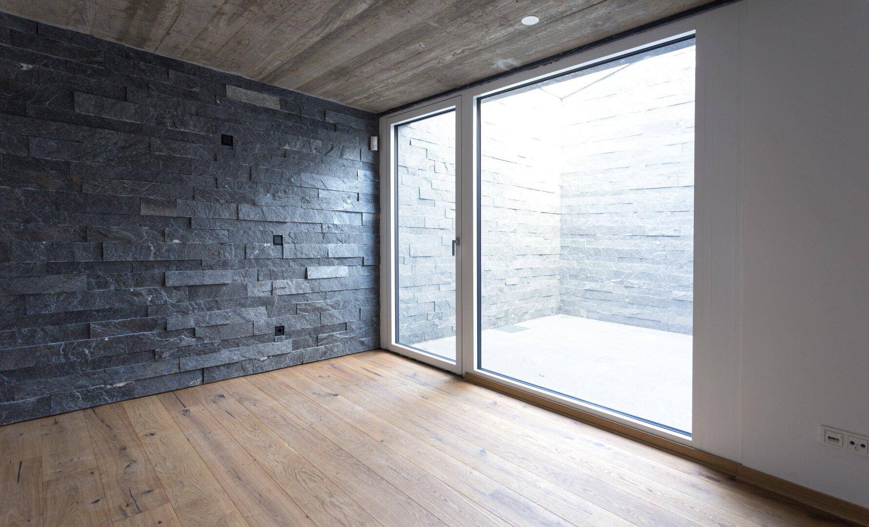 Heller Raum mit Holzboden, Natursteinwand und großer Fensterfront zu einem Innenhof.