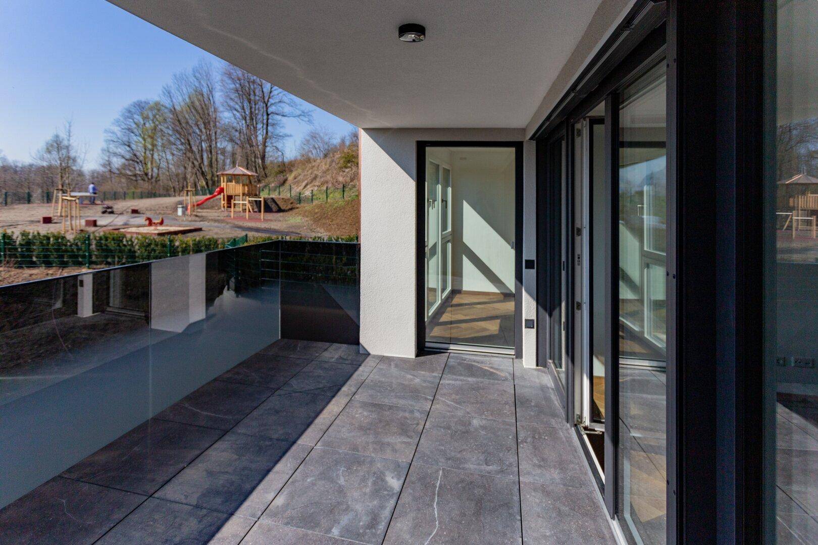 Geräumiger Balkon mit Fliesenboden und Glasbrüstung, mit Blick auf den Spielplatz.