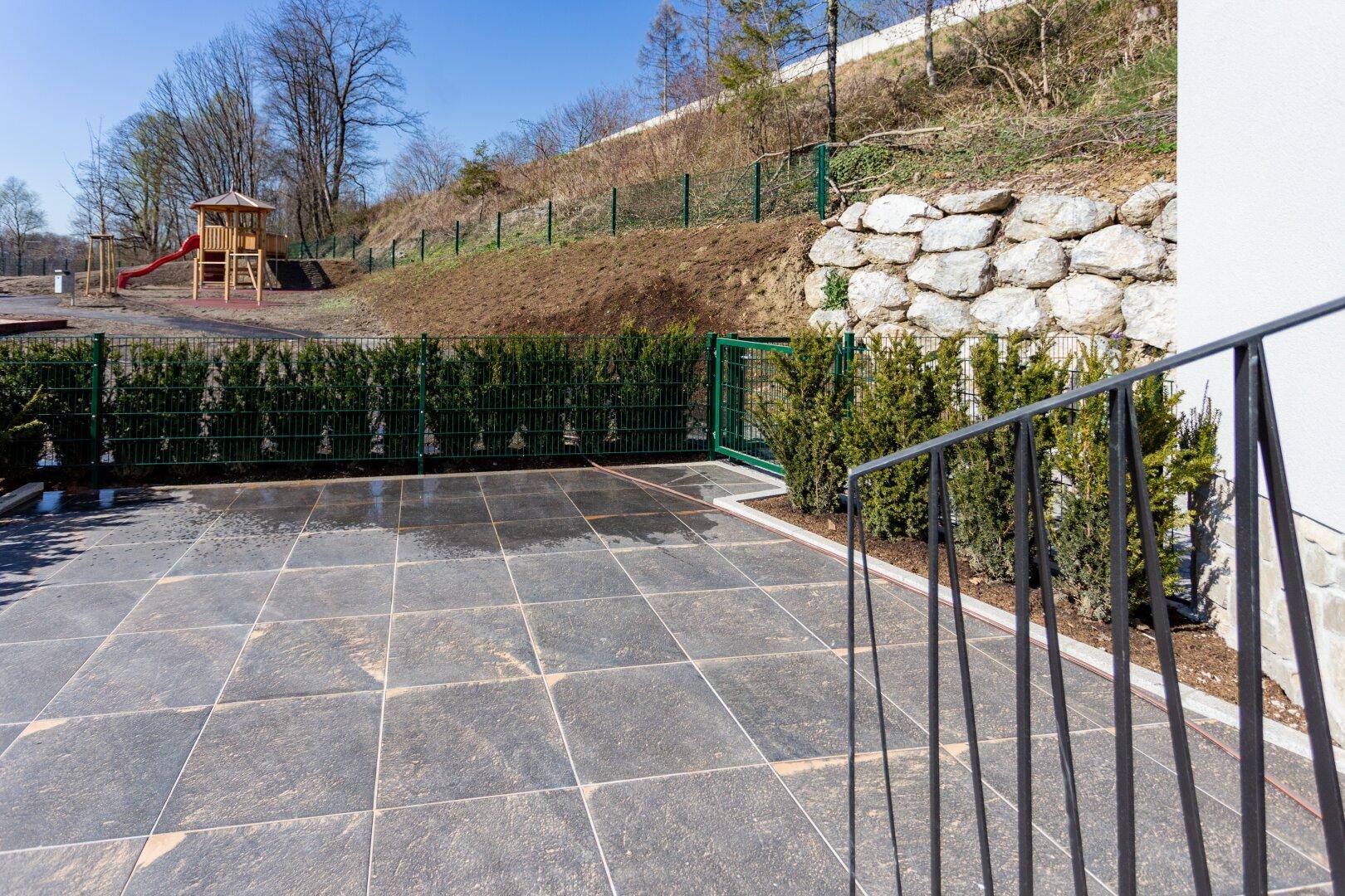 Große gepflasterte Terrasse mit Blick auf einen Spielplatz und eine Natursteinmauer.