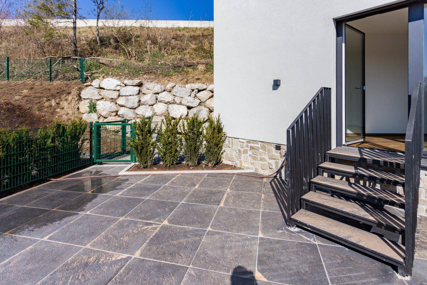 Gepflasterte Terrasse mit Treppe, Natursteinmauer und grünen Büschen.