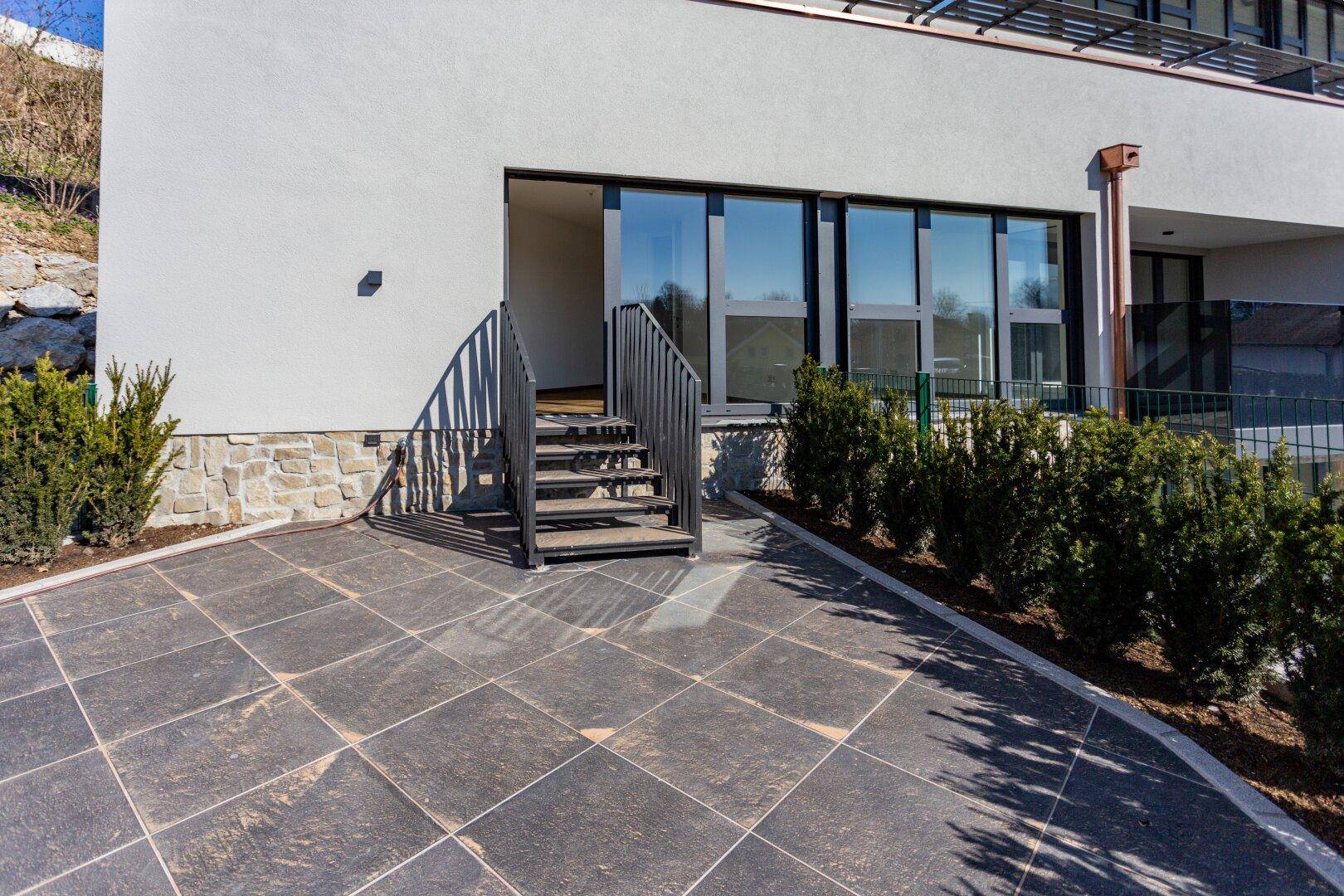 Große gepflasterte Terrasse vor dem Gebäude mit Treppe und gepflegten Hecken.