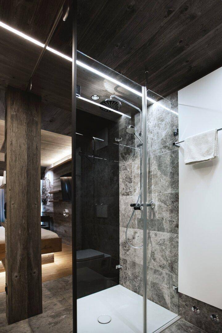 Modernes Badezimmer mit einer begehbaren Dusche, grauen Fliesen und einer Holzsäule, ergänzt durch Deckenbeleuchtung.