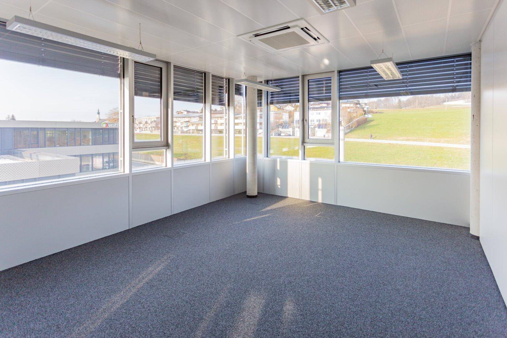 Leeres, helles Büro mit großen Fenstern, Teppichboden und weitem Blick ins Grüne.