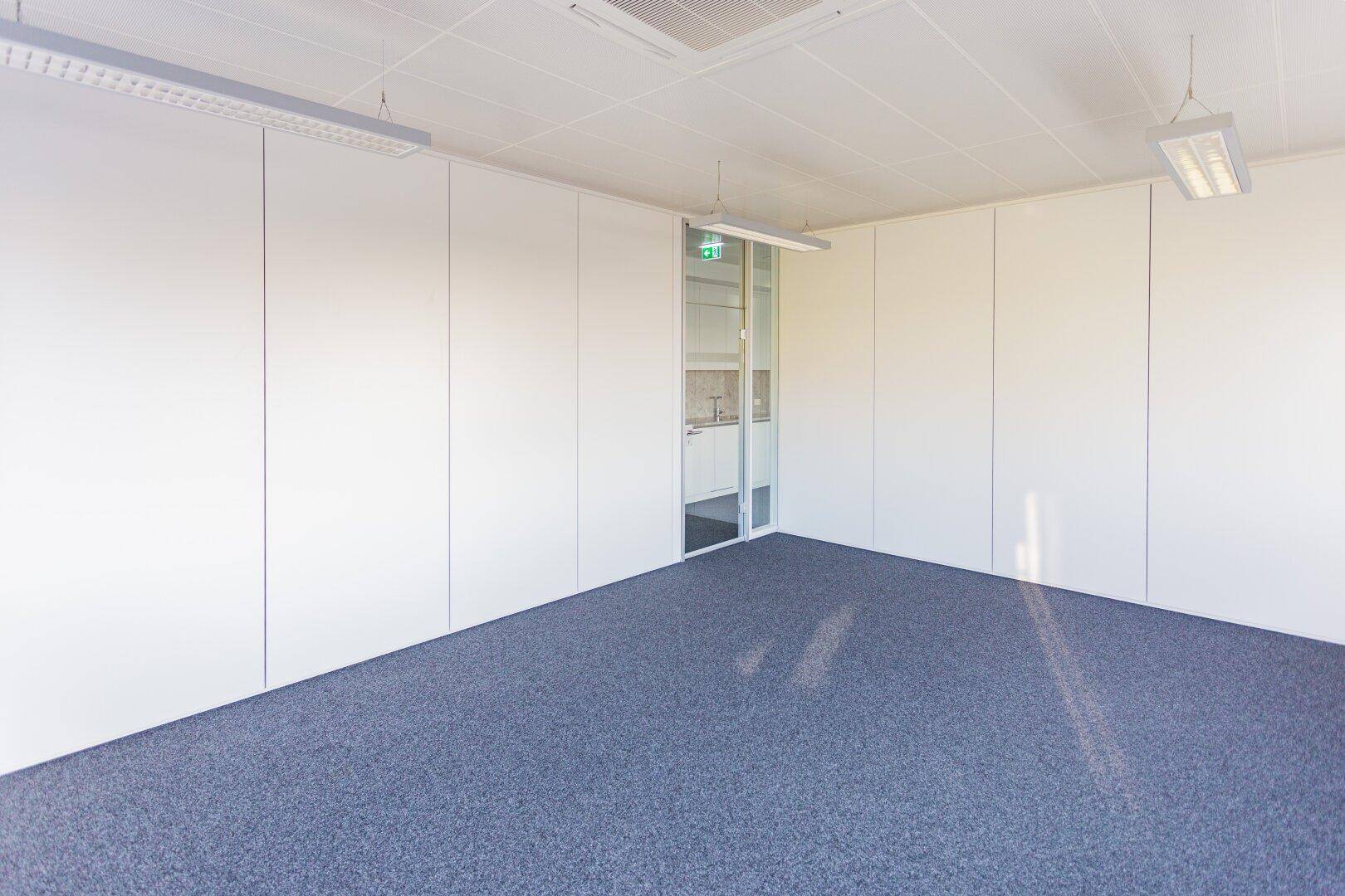 Leeres Büro mit Teppichboden, weißen Wänden und einer Glastür zu einem Nebenraum.