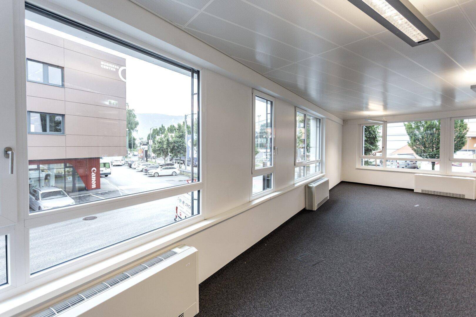Büro mit Teppichboden und großen Fenstern, die einen Blick auf die Straße und umliegende Gebäude bieten.