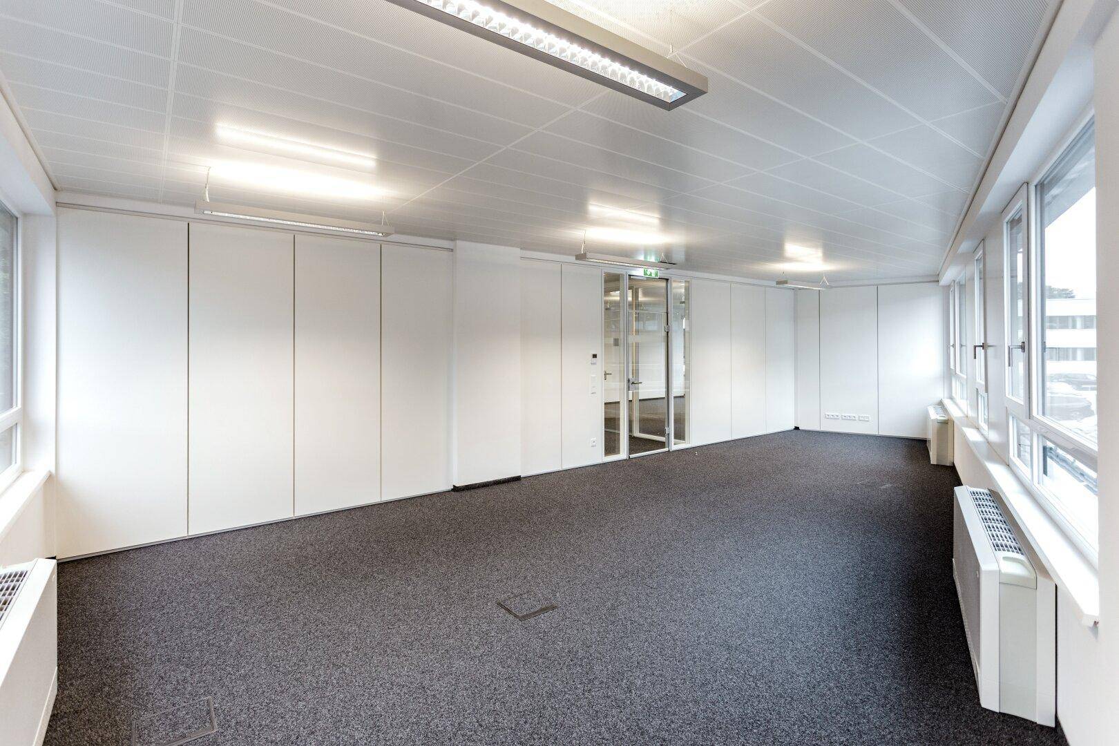 Leeres Büro mit Teppichboden, weißen Wänden und einer Glaswand, die den Raum hell und offen wirken lässt.