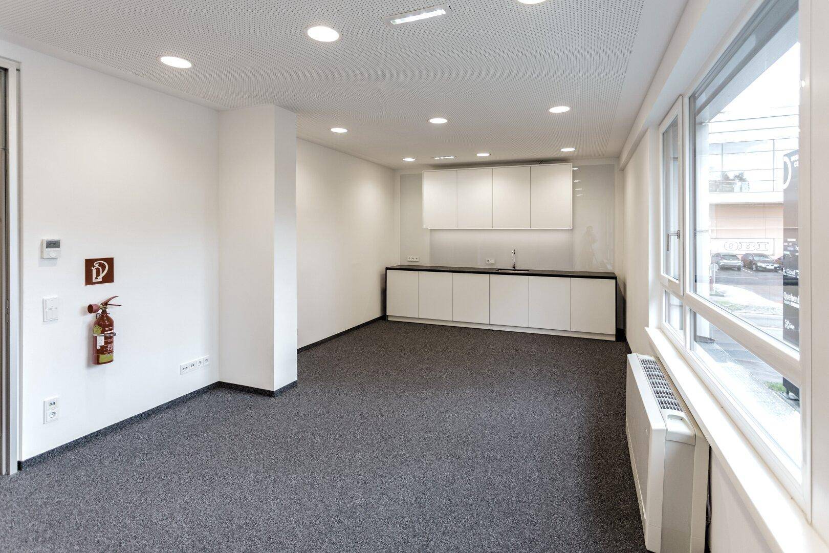 Moderne Teeküche in einem Büro mit weißen Schränken, Spüle und viel Tageslicht.