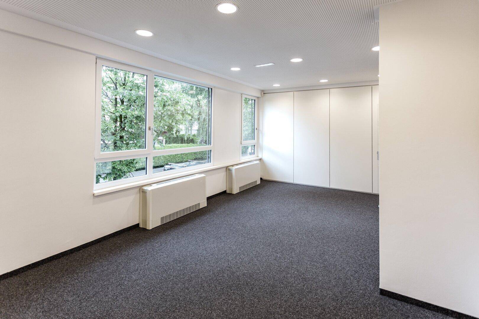 Leeres Büro mit Teppichboden und großen Fenstern, die einen Blick auf die grüne Umgebung bieten.