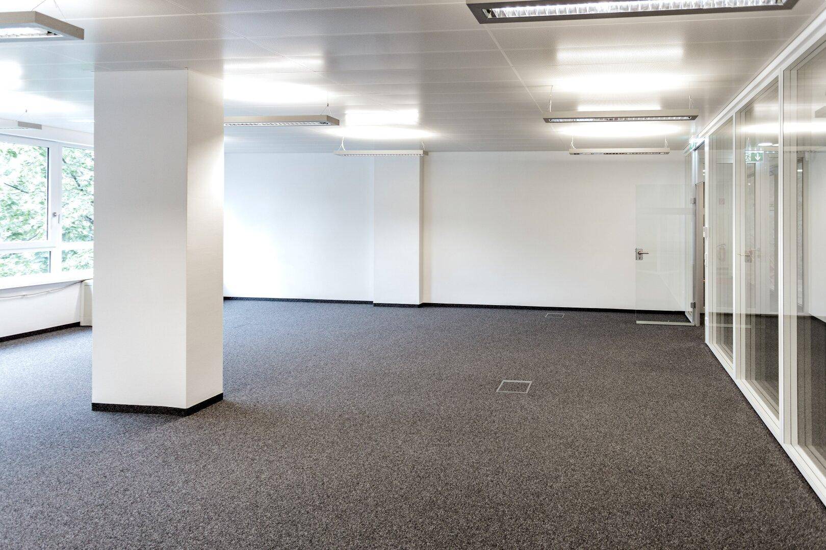 Geräumiges Büro mit Teppichboden, Säulen und großen Fenstern, die den Raum hell und offen gestalten.