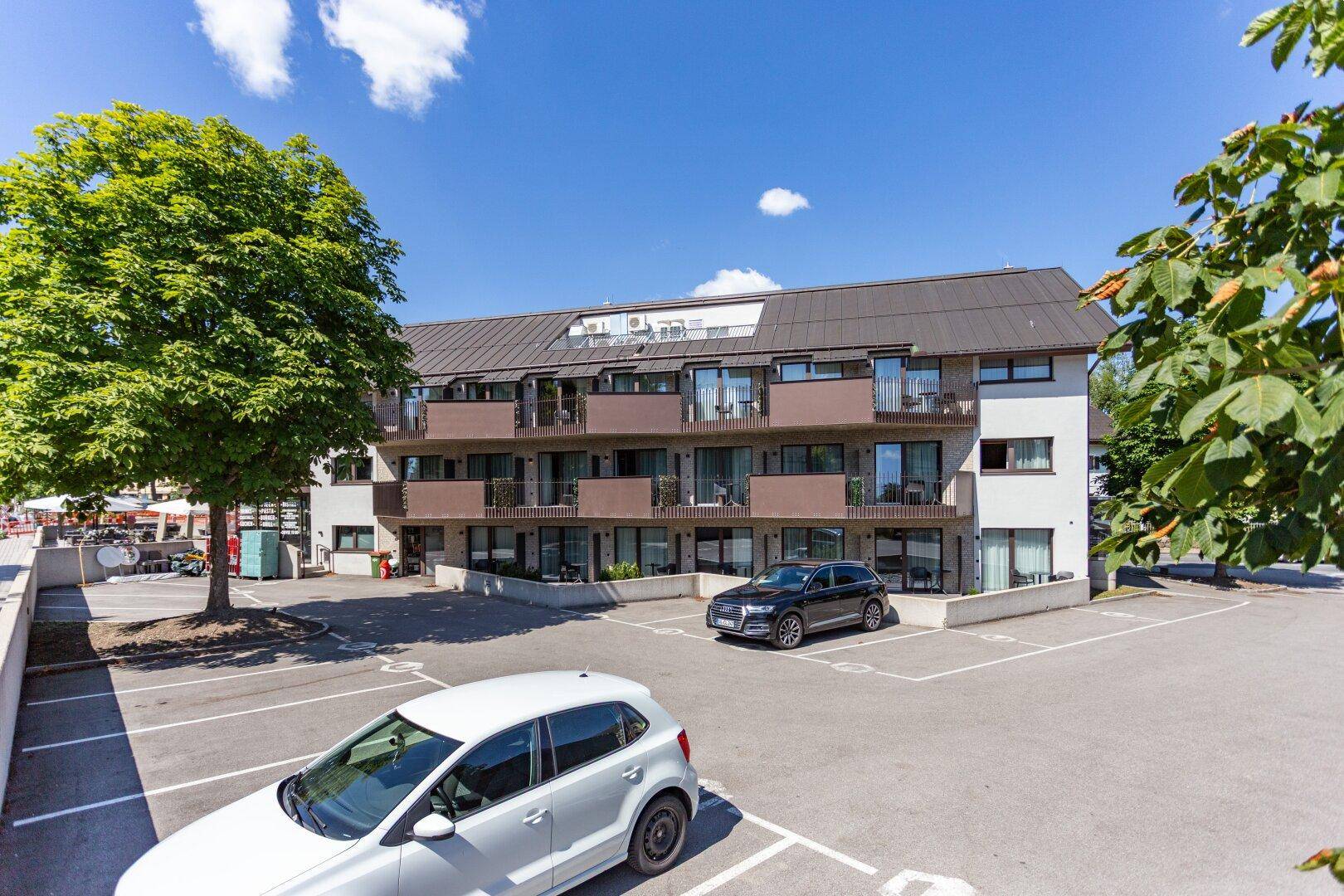 Mehrstöckiges Gebäude mit zahlreichen Balkonen und einem davorliegenden Parkplatz unter blauem Himmel.