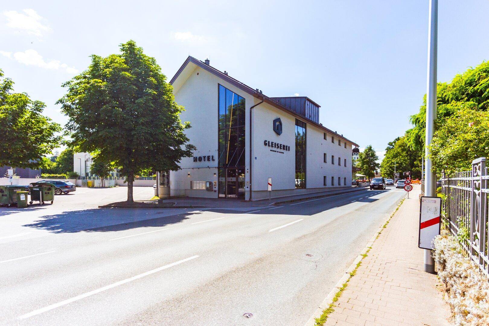 Außenansicht eines modernen Gebäudes mit großer Glasfassade und dem Schriftzug "Hotel", an einer Straße gelegen.