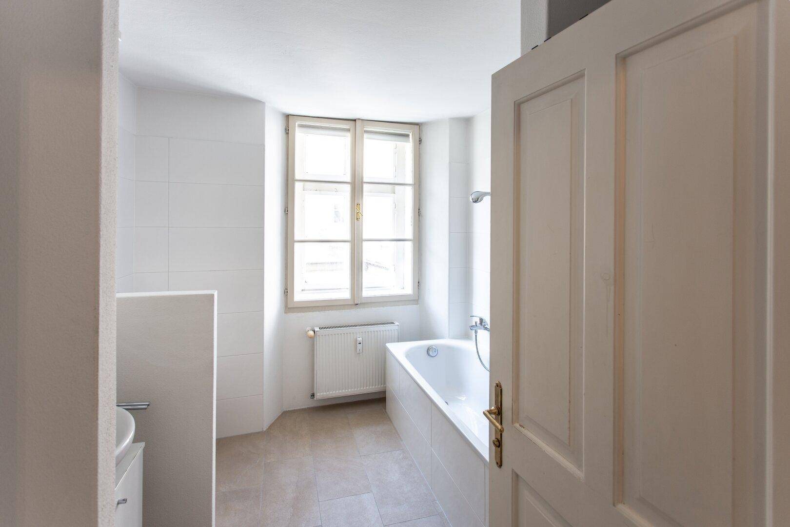 Helles Badezimmer mit Badewanne, Fenster und weißen Fliesen, bereit zur Modernisierung.