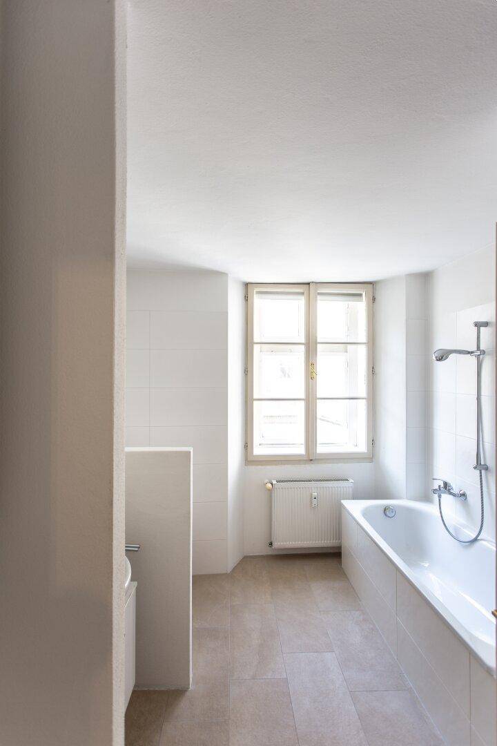 Helles Badezimmer mit Badewanne, Fenster und weißen Fliesen, mit Platz für einen Waschtisch.