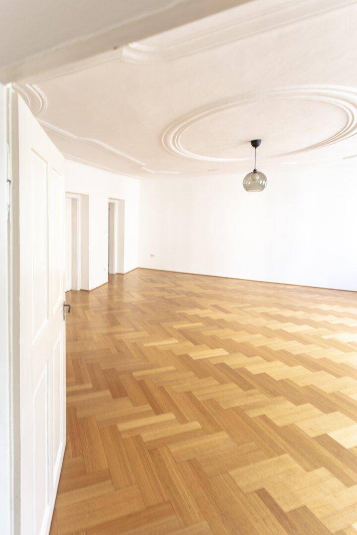 Großes, helles Zimmer mit Fischgrätparkett und eleganter Stuckdecke, mit Zugang zu weiteren Räumen.