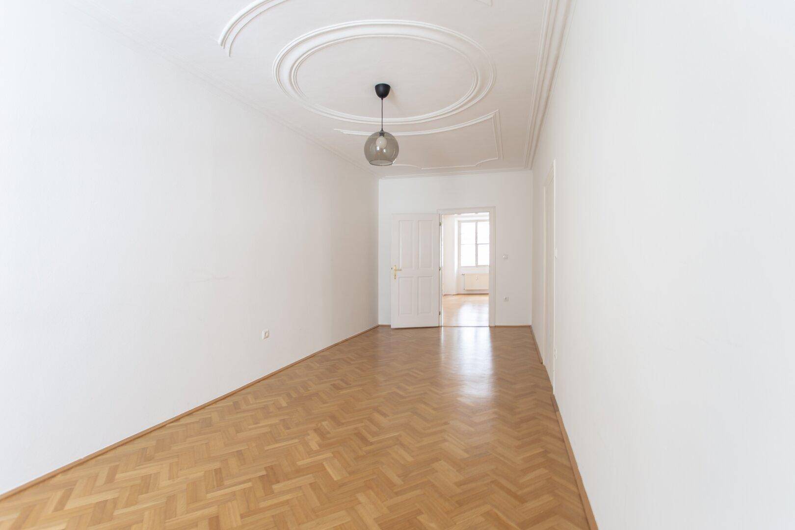 Geräumiges, helles Zimmer mit Fischgrätparkett und Stuckdecke, mit Zugang zu einem weiteren Raum.